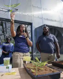 A woman holds a plant with roots while speaking into a microphone. Two people stand nearby, with one holding a jar of seedlings. In the background, greenhouse equipment is visible.
