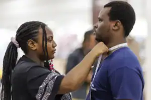 A woman takes measurements around a man's neck using a measuring tape in a clothing store. Both individuals are focused on the task.