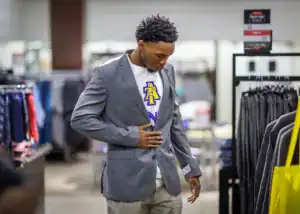 A young man fittings a gray blazer over a T-shirt while shopping in a clothing store. He is focusing on the fit, surrounded by racks of formal attire.