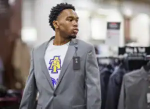 A young man wearing a gray blazer over a white T-shirt stands in a clothing store. He has short, curly hair and is looking to the side, with a price tag still attached to the blazer.