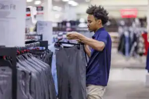 A young man examines a pair of gray pants on a hanger while shopping in a clothing store with racks of merchandise in the background.
