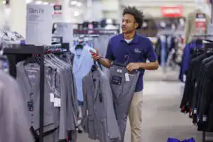 A young man in a store holding a gray suit jacket and pants, looking at clothing displays. He is dressed in a blue polo shirt and appears to be assisting customers or shopping.