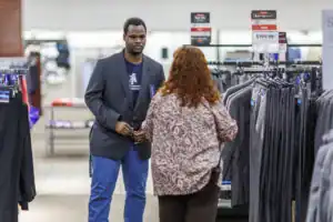 A man in a blazer stands in a clothing store, facing a woman with curly hair, who is looking at a rack of suits. Sale signs are visible in the background.
