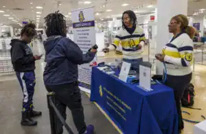 A career services booth features two staff members in striped sweaters, assisting students. One student is on their phone, while another interacts with the staff, discussing services and resources available.