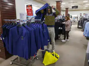 A person in a striped shirt tries on a blue suit jacket while standing in a clothing store. Another shopper browses through items nearby. A yellow bag lies on the floor.