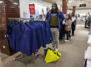 A person in a blue blazer and striped shirt stands near a rack of blue blazers in a store. A yellow bag is on the floor nearby. Another shopper browses in the background.