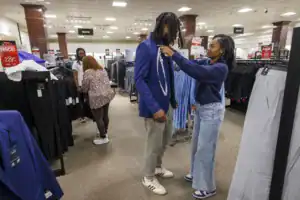 A man in a blue blazer stands while a woman fits him with a measuring tape in a clothing store. Another woman shops in the background among racks of pants.