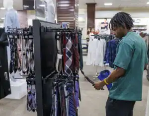 A person examines a selection of ties displayed on a rack in a clothing store, holding a blue shirt in one hand while looking closely at the ties.