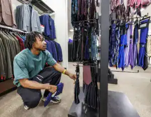 A person with dreadlocks squats in a clothing store, examining ties on a display rack. Men's suits are visible in the background.