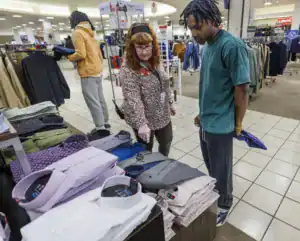 A woman points to dress shirts on a table while a man looks on, holding a blue item. Clothing racks are visible in the background.