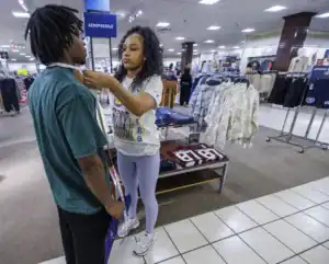 A woman in a store measures a man's neck with a tape measure. Shelves displaying clothing are visible in the background.