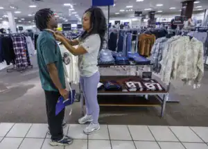 A woman measures the neck of a man in a clothing store. Clothing racks with various shirts and jeans are visible in the background. The scene suggests they are selecting or trying on clothes together.