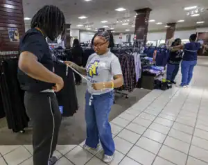 A young woman in glasses measures the waist of a young man in a clothing store. They are surrounded by racks of clothing, while others shop in the background.