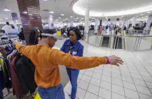 A young man with headphones stands with arms outstretched, wearing a measuring tape around his chest. A woman in glasses and denim looks at him, both in a clothing store surrounded by racks of apparel.