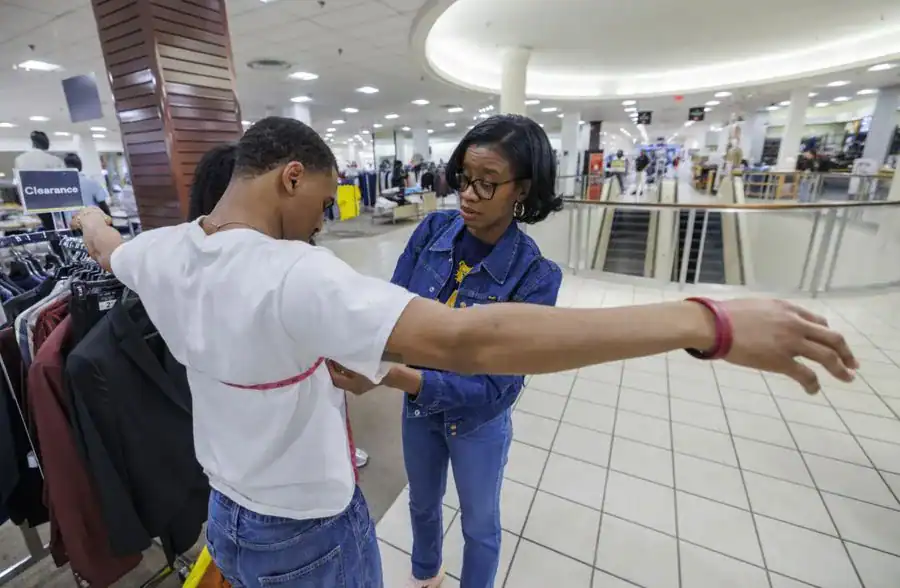 A woman measures a man's chest while shopping in a clothing store, surrounded by racks of apparel and shoppers in the background.