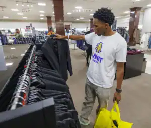 A young man examines a black suit jacket on a hanger while holding yellow shopping bags in a clothing store. Rows of suits and formal wear are visible in the background.