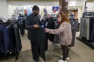 A man in a black suit and woman in a patterned top are shopping together in a clothing store, examining a pair of pants while discussing options among several suit jackets on display.