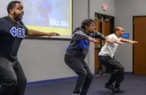 Three young men perform synchronized squat movements in a classroom setting, demonstrating energy and focus. A colorful "ROLL CALL" slide is visible in the background.