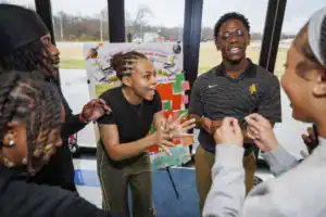 A group of five young adults interact enthusiastically in a bright room, engaged in discussion. A colorful poster is displayed in the background, suggesting a collaborative activity or event.