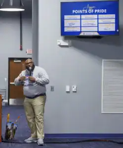 A speaker in a gray sweatshirt presents in a classroom. Behind him is a screen displaying "Points of Pride" with various statistics about a university. A bag is placed on the floor beside the speaker.