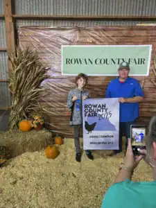 A young contestant holds a blue ribbon and a sign declaring them the Grand Champion for a broiler pen at the Rowan County Fair 2025, while an adult poses beside them. Pumpkins are arranged in the background.
