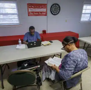 A man works on a laptop at a table, while a woman sits nearby reading a book. The room features a red tablecloth and a sign related to a church.