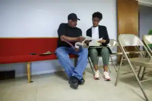 A man and a woman sit together on a red bench, reviewing documents. The man wears a black cap and t-shirt, while the woman sports glasses and a black jacket. Metal chairs are positioned nearby against a light gray wall.