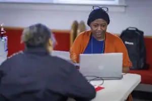 A woman in an orange shawl sits at a table working on a laptop, focused on her task. Another person, viewed from behind, is also at the table. In the background, there’s a black backpack and a red wall.