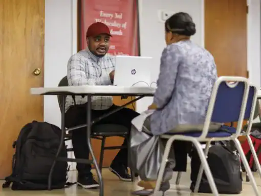 A man in a cap and plaid shirt sits at a table facing a woman in a gray outfit, both engaged in conversation. A laptop is open in front of them, with bags nearby.