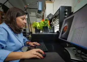 A woman in a lab coat works at a computer, analyzing data. Potted plants are visible in the background, with equipment arranged on the desk around her.