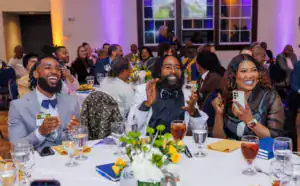 Three smiling people at a banquet table clap and celebrate. The man in a light suit and bow tie, the man in a tuxedo, and the woman with a phone all express joy, surrounded by guests and floral centerpieces.