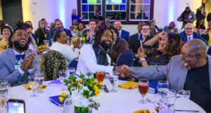 A diverse group of people at a celebratory event enjoy laughter and conversation. Two men shake hands while smiling, with others applauding and engaging. Tables are set with drinks and flowers.