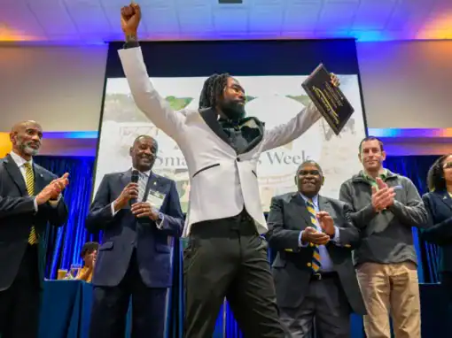 A man in formal attire triumphantly raises a plaque while standing on stage. He is surrounded by five applauding people, including men in suits and women in business attire, with a colorful backdrop and a presentation screen behind them.