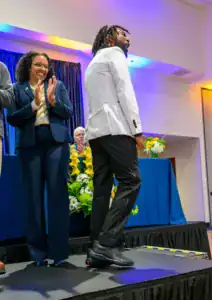 A man in a white blazer celebrates on stage, while a woman in a navy suit applauds. In the background, a man sits at a table with flowers and colorful lighting enhances the event's atmosphere.