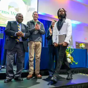 A diverse group of four people stands on stage, smiling and clapping. One man in formal attire confidently walks past, holding a clipboard. A colorful backdrop features flowers and a logo for a small farm initiative.
