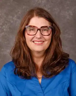 Smiling woman with long brown hair wearing colorful glasses and a blue shirt, set against a gray background.