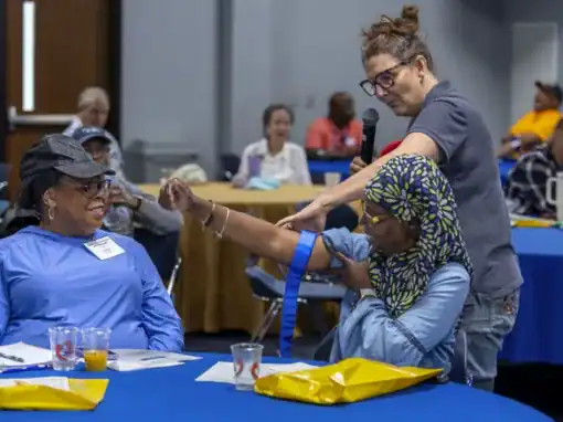 A woman demonstrates a procedure on another woman's arm at a workshop, while an audience observes. Tables are set up with blue tablecloths, and some attendees are engaged in conversation.