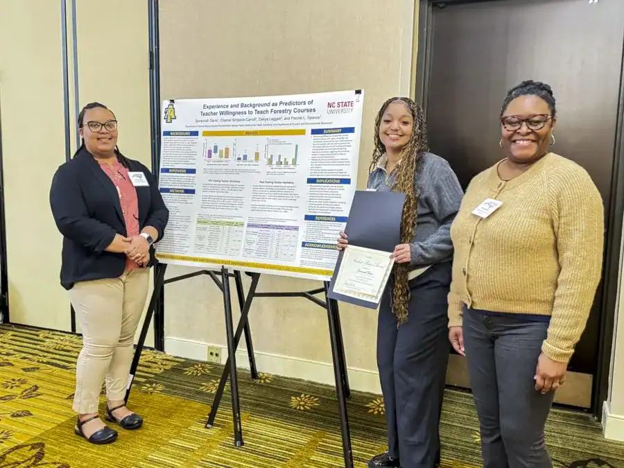 Three women stand in front of a research poster on teaching forestry courses. One holds a certificate, while the others smile at the camera. The background features a neutral-colored wall and a patterned floor.