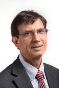 A man with short, dark hair and glasses wears a gray blazer and a striped tie. He is facing forward and smiling slightly against a light background.