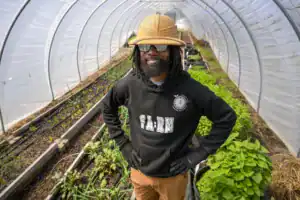 Man smiling in a greenhouse, wearing a straw hat and sunglasses. He stands confidently among rows of growing plants, dressed in a black hoodie and brown pants.