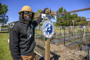 Smiling man with dreadlocks and sunglasses stands next to a sign that reads "The McKoy Way" and features the "Homegrown By Heroes" logo in a community garden setting.