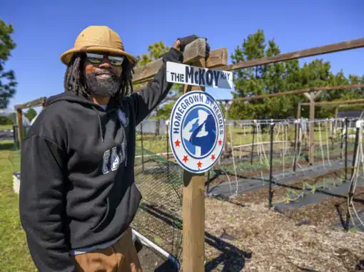 Smiling man with dreadlocks and sunglasses stands next to a sign that reads "The McKoy Way" and features the "Homegrown By Heroes" logo in a community garden setting.