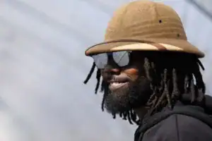A smiling man with dreadlocks wears a straw hat and sunglasses, standing against a blurred background.