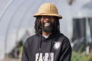 A man with long dreadlocks and a wide smile wears a beige hat and a black hoodie with "FARM" printed on it, standing in a greenhouse with green plants in the background.