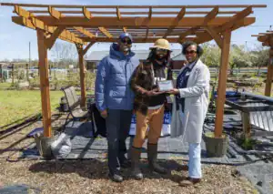 Three people stand outdoors under a wooden structure in a garden setting. One person holds an award while the others smile. A chair and various garden elements are visible in the background.