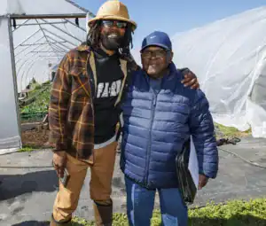 Two men pose together outdoors in a farm setting, smiling. One has long dreadlocks and wears sunglasses and a plaid shirt, while the other wears glasses and a blue puffer jacket, holding a folder. A greenhouse is visible in the background.