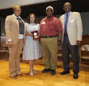 Four individuals pose together, holding a clock award. The group stands in a warmly lit room, with chairs in the background. They are dressed in formal attire and display cheerful expressions.