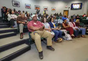 A diverse audience is seated in a lecture hall, attentively listening to a speaker. Some individuals are taking notes or using devices. The walls feature framed photographs and a digital presentation is displayed.