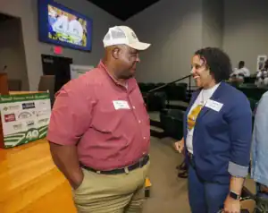 Two attendees engage in conversation at an event. A man in a red shirt and cap listens while a woman in a blue jacket smiles and gestures, showcasing an enthusiastic exchange. A screen in the background displays "Small Farms Week.