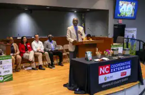 A speaker addresses an audience at a podium during an event. Four panelists sit nearby, with a table displaying promotional materials and flowers. A presentation on the screen highlights "Small Farms Week.